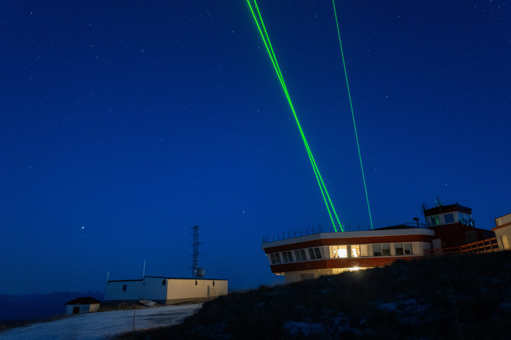 Photo of the Alomar observatory at night. Stars in background, green laser beams shooting upwards.