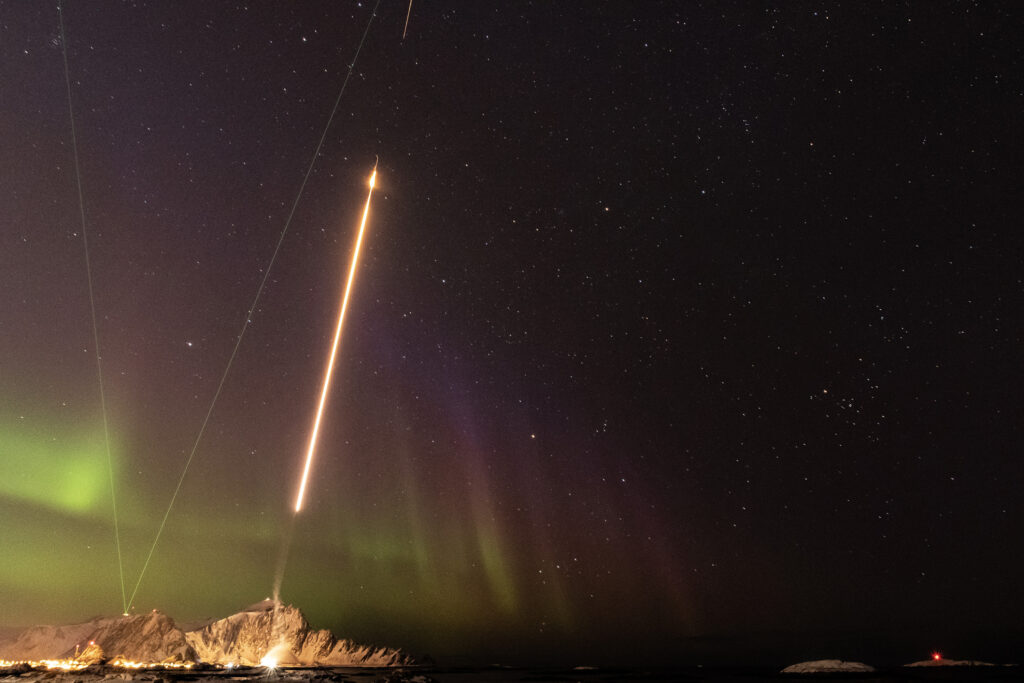 Long-exposure photo of a rocket launch, with aurora and start in the background.